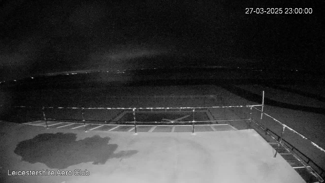 A dark webcam image showing a mostly empty outdoor area. In the foreground, there is a flat surface, likely a runway or platform, with a railing along the edge. In the background, faint outlines of distant lights can be seen, suggesting a hazy horizon. The overall scene is dim, indicating nighttime, with no discernible features in the sky or surrounding area. The date and time displayed in the corner indicate it's March 27, 2025, at 11:00 PM.