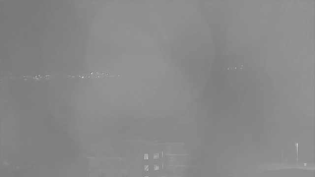 A grayscale image with a blurred view showing faint lights in the distance, likely from buildings or streetlights. The foreground features dark shapes that may represent structures or trees, but they are not clearly defined due to the lack of visibility. Overall, the scene has a foggy or misty appearance, making details hard to discern.