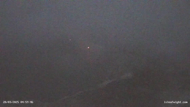 A dark, foggy scene captured by a webcam, with very limited visibility. A few faint lights can be seen in the distance, likely from a shoreline or distant landmarks, surrounded by an indistinct, blurred background. The overall atmosphere feels eerie and quiet. The timestamp at the bottom indicates the date and time of the image.