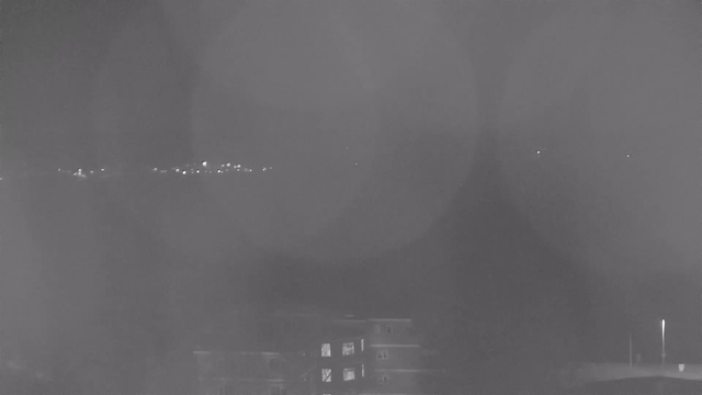 A dimly lit scene showing a dark expanse of water with faint lights from a distant shore. The overall image has a monochrome quality, with circular light reflections appearing prominently throughout. In the foreground, silhouettes of buildings can be discerned, some with lights in their windows. The atmosphere feels quiet and serene, with a hazy appearance due to the lighting.
