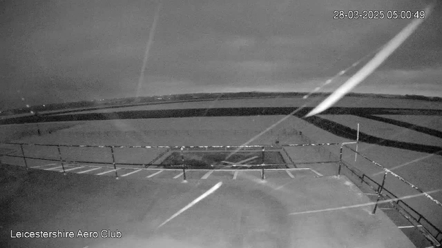 A dark, nighttime scene from a webcam at the Leicestershire Aero Club. The image shows a tarmac area with white striping, likely a parking lot, in the foreground. Beyond the lot, there is an open field with a low horizon line partially obscured by clouds. The scene is illuminated by faint light, suggesting early morning, with streaks of light cutting diagonally across the image.