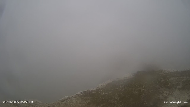 A foggy scene with a greyish sky and indistinct visibility. The ground is uneven with some rocky surfaces and grass visible in the foreground. The atmosphere appears damp and lacks clear outlines or distinct features.