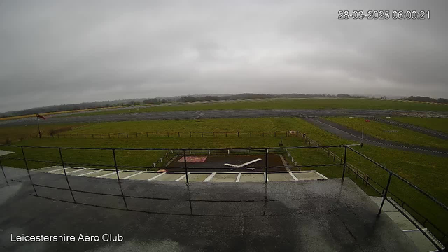 A cloudy sky with low visibility covers a landscape view from a webcam at the Leicestershire Aero Club. In the foreground, there is a railing and platform. Below, a grassy area leads to a runway marked with a white X. In the distance, a patch of trees is visible along the horizon, while a windsock indicating wind direction is present on the left. The overall atmosphere is quiet and overcast.