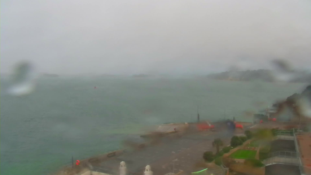 A hazy, overcast view of a coastal area during rainy weather. The foreground shows a waterfront with a few structures and paths, along with some greenery. The water appears choppy and gray, and there are faint outlines of distant islands or land masses visible through the fog. The scene is drenched in a dull light, with droplets of water on the lens partially obscuring the view.