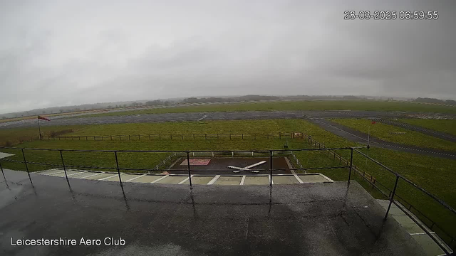 A cloudy and overcast scene at Leicestershire Aero Club. In the foreground, there is a gray pavement area with a railing. The ground is wet, suggesting recent rain. A single small airplane with a white body and wings is visible on the ground, parked on a designated area. In the background, the runway and a large grassy field extend into the distance, surrounded by a wooden fence. A windsock indicates the wind direction. The overall atmosphere is somber due to the gray sky. The time displayed is 06:59:55 on March 28, 2025.