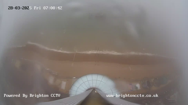 A cloudy, foggy view from a high vantage point, showing a sandy beach and gentle waves lapping at the shore below. In the foreground, the curved edge of a structure is visible. The date and time display is in the upper left corner, and text indicating the source of the webcam appears in the lower left and right corners.