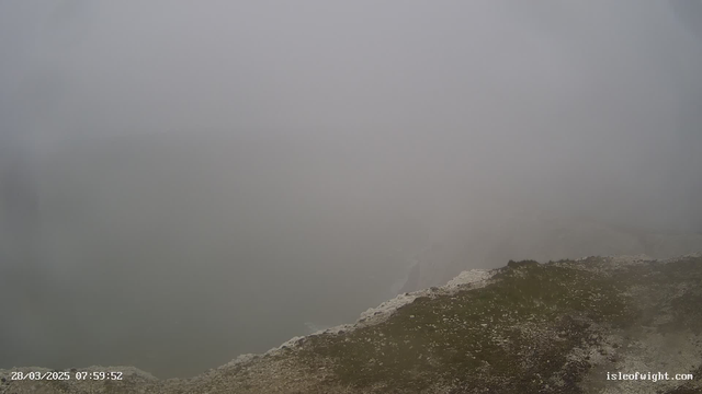 A cloudy and foggy scene is depicted with visibility nearly obscured. There are hints of land and water, but details are indistinct due to the heavy mist. Grass and rock textures are visible in the foreground, while the horizon fades into the gray fog. The time-stamp indicates it is early morning.