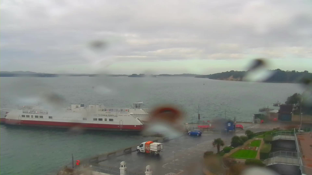 A gray, overcast view of a harbor with a large ferry boat docked at the edge of the water. The ferry has a white body and red lower sections, and signs visible on the side. In the foreground, there is a truck parked on the wet pavement, with a blue car nearby. Various greenery and palm trees are planted along the walkway next to the harbor, which is surrounded by distant hills and a cloudy sky. Raindrops are visible on the camera lens, causing blurriness.