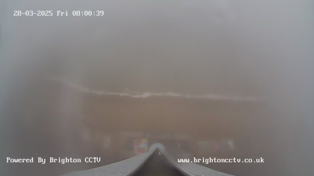 A foggy and blurred view from a webcam, showing a sandy beach with some faint outlines of water. The top of the image displays the date and time: "28-03-2025 Fri 08:00:39." The bottom of the image includes the text "Powered By Brighton CCTV" and a website URL.