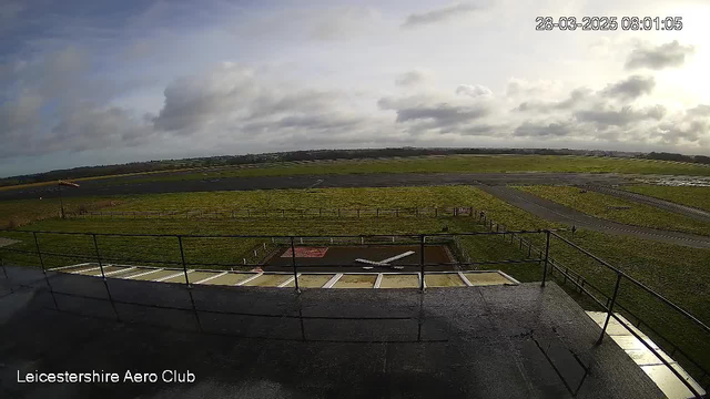 A view of a rural airfield from an elevated perspective. The foreground includes a fenced area with a painted white aircraft on the ground. The airfield is surrounded by green grass and a clear blue sky with scattered clouds. In the distance, rolling hills and more open fields are visible, creating a sense of openness and rural landscape. The image is timestamped with the date and time at the top right corner.