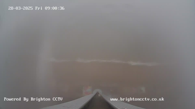 A foggy view of a beach from a webcam. The foreground shows a wide walkway or pier leading towards the beach. The water is barely visible due to the dense fog, with some soft waves and a sandy shore in the background. The sky is grayish, adding to the overall misty atmosphere. The date and time are displayed at the top, indicating March 28, 2025, at 09:00:36.