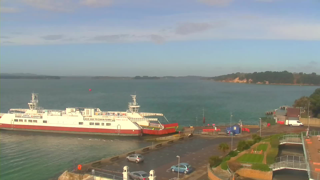 A white and red ferry is docked at a port, with the calm blue water of a harbor surrounding it. In the background, there are green hills and a sandy shore, with some trees lining the edge. Near the ferry, there is a parking area with a few cars and a van, along with several colorful barriers and a small blue building. The sky is mostly clear with a few scattered clouds.