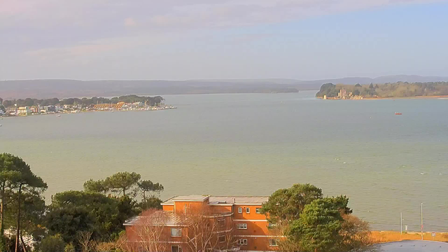 A tranquil view of a body of water surrounded by trees and buildings. In the foreground, an orange-colored building with multiple windows and some bare trees. To the left, there is a marina with several small boats and colorful buildings lining the shore. In the distance, a peninsula with greenery and a structure that resembles a castle is visible. The sky is mostly clear with soft clouds, and the water reflects a calm, light blue and green hue.
