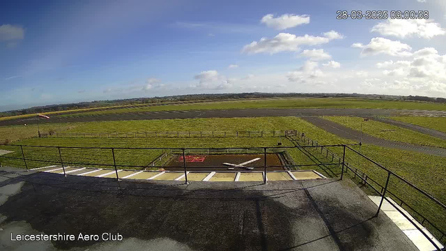 A view from a webcam at Leicestershire Aero Club shows a wide open field under a clear blue sky. The horizon is lined with distant hills. In the foreground, there is a black surface with a white airplane silhouette on it, possibly a helipad or landing area. A grassy area with a fence is visible to the left, and a winding runway stretches across the center of the image. Several clouds are scattered across the sky, and the time stamp in the top right corner indicates the image was taken on March 28, 2025, at 09:00:58.