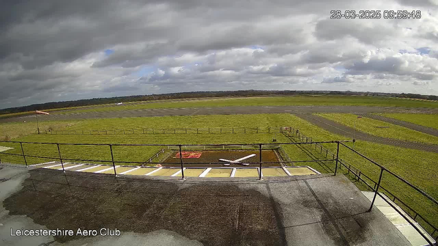 A view from a webcam at Leicestershire Aero Club, showing a large, open grassy area with a runway. The sky is filled with clouds, indicating potential overcast weather. There are several light poles and a windsock blowing softly to the left. A small airplane is parked on the runway, and in the foreground, there's a flat area with some markings and a model airplane. Fences border the grassy field, and the ground appears damp.