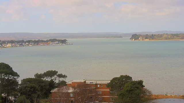 A scenic view of a coastline featuring calm waters of a bay. In the foreground, there is a building with multiple windows surrounded by trees. To the left, a marina is visible with several boats docked. The horizon includes green hills under a partially cloudy sky. On the right, there is a small island with a building at its shore. The overall atmosphere suggests a peaceful, natural setting.