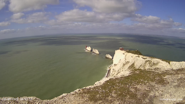 A panoramic view of a coastline with white chalk cliffs dropping into the greenish-blue sea. In the distance, two rocky formations with bright red lighthouses are visible, positioned on the left side of the image. The sky is partly cloudy with patches of blue, and the water shows gentle ripples reflecting light. The foreground features grassy land sloping down towards the water.