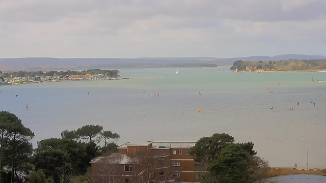 A scenic view over a body of water, with a mix of calm and slightly choppy waves. In the foreground, there are trees and a brown building, partially visible. The water features various sailboats, windsurfers, and colorful markers. The background shows a shoreline with scattered buildings and a distant green landscape, including tree-covered hills and a small island with a structure. The sky is partly cloudy, creating a soft light across the scene.