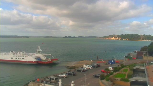 A view of a harbor on a cloudy day. A large white and red ferry is docked along the shore, with a few small boats visible in the water. The background features a shoreline with trees and cliffs. In the foreground, there is a parking lot filled with cars, including a few red vehicles. The sea has varying shades of blue and green, with gentle waves.