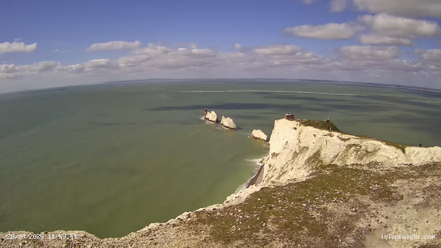 A panoramic view of a coastal landscape featuring striking white chalk cliffs along the shoreline. The sea is a greenish-blue color, with gentle waves visible. Small rocky outcrops emerge from the water, and there are two red lighthouses positioned on different rocks. The sky above is bright blue, dotted with fluffy white clouds. The foreground includes a rocky terrain, leading to the edge of the cliffs. The scene captures a sunny day with clear visibility.