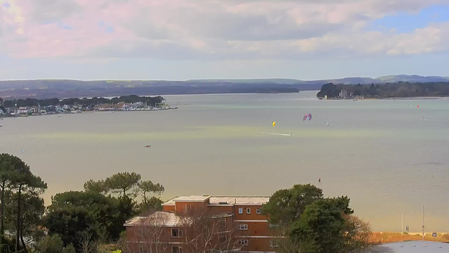 A serene view of a large body of water with gentle waves, bordered by distant hills. On the left, a small marina is visible with several boats docked. In the foreground, there are patches of greenery and trees. In the water, small sailboats and kite surfers are seen, with colorful kites in shades of yellow and pink. The sky is partly cloudy with soft pastel colors, and the scene conveys a calm, outdoor atmosphere.