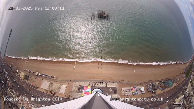 An aerial view of the coastline showing a sandy beach with waves gently lapping at the shore. A pier is visible extending into the water, with an abandoned structure sitting offshore. The scene includes various colorful attractions and structures along the beach. The image captures a clear blue sky with some scattered clouds overhead. The date and time are displayed at the top of the image, indicating it is March 28, 2025, at noon.
