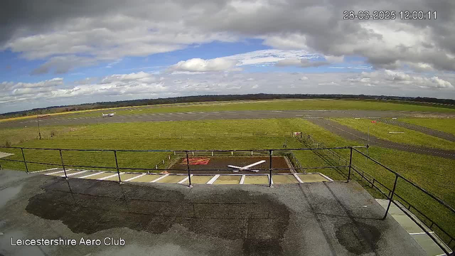 A panoramic view of a grassy airstrip at Leicestershire Aero Club under a mostly cloudy sky. In the foreground, there is a railing and a small runway area with a white cross marking. In the background, a small aircraft is seen taxiing on the airstrip. The landscape is flat, with fields stretching towards the horizon.