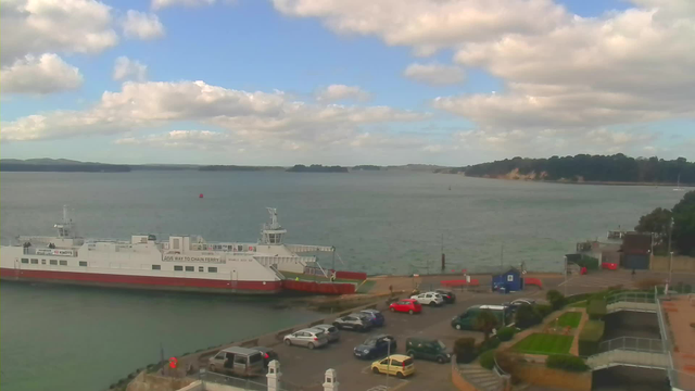 A view of a coastal area with a large ferry docked at the shore. The ferry is primarily white with a red bottom and has visible signage. The water is calm with distant hills and islands in the background. Fluffy clouds are present in a blue sky. In front of the ferry, there is a parking lot with several cars, including red, green, blue, and yellow vehicles. The shoreline features some greenery and steps leading up from the water's edge.