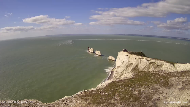 A scenic view of cliffs along a coastline under a bright blue sky with scattered clouds. The cliffs are predominantly white and rocky, leading down to a greenish sea. In the distance, there are a few rocky outcrops protruding from the water, with some buildings on top. The shoreline is partially visible, with a small beach area. The scene conveys a peaceful and natural landscape with the ocean gently lapping at the shore.