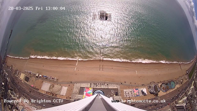 Aerial view of a beach and ocean under bright sunlight. The sandy shore stretches along the bottom, with small figures of people walking and scattered objects visible. Waves gently lap against the beach, and a pier extends into the ocean at the center. The water reflects sunlight, creating a shimmering effect. At the bottom of the image, there are colorful beach huts and a large building with a circular structure, possibly an amusement area. The date and time are displayed in the top left corner.
