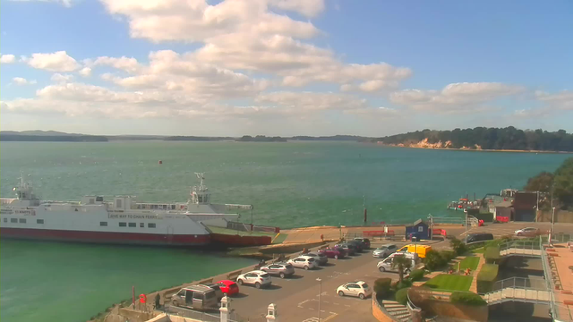 A view of a coastal area with a blue-green body of water. A large ferry boat is docked at the pier, which is surrounded by parked cars. The parking area includes several vehicles of different colors. In the background, there are cliffs and greenery along the shoreline under a partly cloudy sky. The scene has a peaceful atmosphere, reflecting a sunny day by the water.