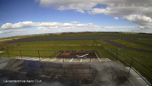 A view from a webcam at Leicestershire Aero Club shows a large green field under a bright blue sky with scattered white clouds. In the foreground, there is a flat rooftop with a railing and a marked area for a helicopter landing, characterized by a large white cross on a dark surface. In the background, a runway is visible with a grassy area and trees lining the far side.