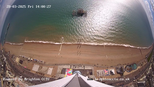 An aerial view of a sandy beach with gentle waves lapping at the shore. Several people are walking along the beach, and there are colorful beach umbrellas and lounge chairs scattered about. In the distance, a pier is visible extending into the water. The scene is framed by a blue sky with a few scattered clouds. At the bottom, there are structures, including a circular pattern and what appears to be an amusement area with rides. The time and date are displayed at the top, indicating it is 14:00 on March 28, 2025.