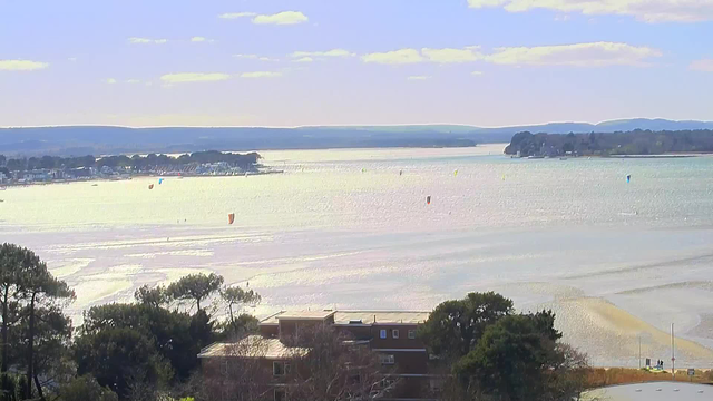 A scenic view of a body of water with gentle waves reflecting sunlight. In the foreground, there are trees and a low building, possibly a coastal structure. The midground features a beach area with a few people visible. In the background, a line of houses runs along the shore. Colorful kites are flying above the water, adding a vibrant touch to the landscape. The horizon showcases rolling hills under a partly cloudy sky.