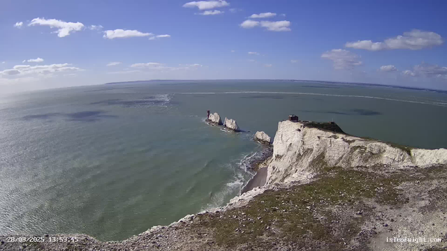 A wide view of the ocean meets the coastline, featuring white chalk cliffs that rise steeply from the water. On the left, there are several jagged rock formations, some with a small lighthouse structure on top. The sea is calm with gentle waves, reflecting light from the sun, while a few scattered clouds float in a bright blue sky. The foreground shows a grassy area with stones, leading to the edge of the cliffs.