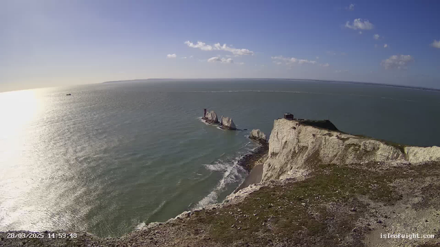A wide view of the coastline featuring white chalk cliffs, with a clear sky and scattered clouds. The sun reflects on the calm sea, creating a shimmering effect. In the distance, several rock formations rise from the water, and a small boat can be seen. The foreground includes grass and rocky terrain leading down to the beach.
