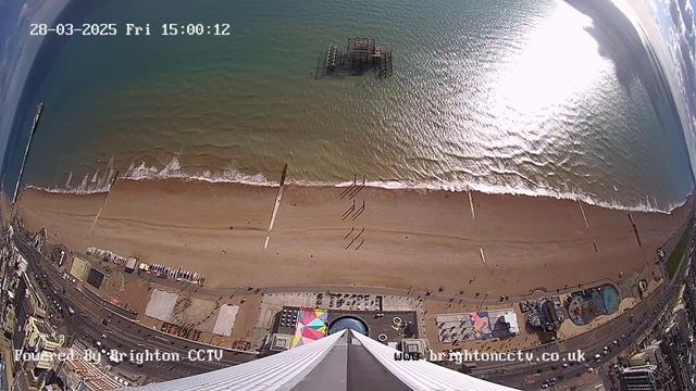 A view from a high angle looking down at a beach with soft sand and gentle waves. Along the shoreline, people are walking, and there are a few piers extending into the water. An old, partially submerged structure is visible in the sea. On the beach, colorful structures and a pool can be seen, along with various amenities and activities. The water reflects sunlight, creating a shimmering effect. The sky is clear with a few clouds. The date and time are displayed at the top of the image.