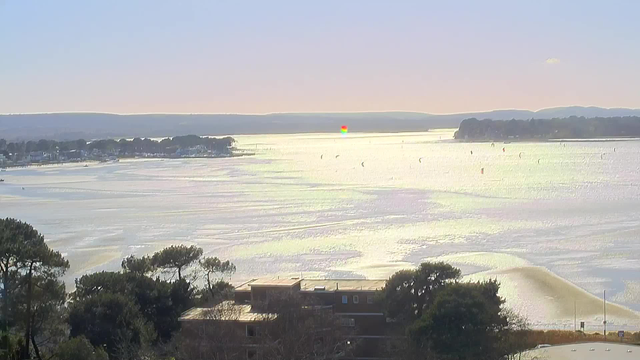 A scenic coastal view featuring a shimmering body of water under a clear sky. In the distance, there are rolling hills and a small forested area. Several colorful kite boarders are seen gliding across the water, and the coastline is lined with houses and trees. The sunlight creates a reflective, shimmering effect on the water's surface.
