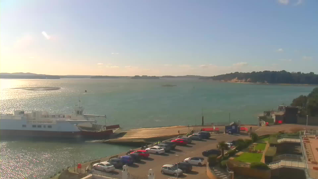A panoramic view of a waterfront scene under a clear blue sky. In the foreground, there is a docked ferry with a white hull, partially obscured by a small ramp. A parking lot is visible, filled with various cars in colors like red and blue. Beyond the ferry, the turquoise water gently ripples reflecting the sunlight. In the background, a distant shoreline features rolling hills and trees, with a sandy beach area. The scene gives a serene coastal atmosphere.