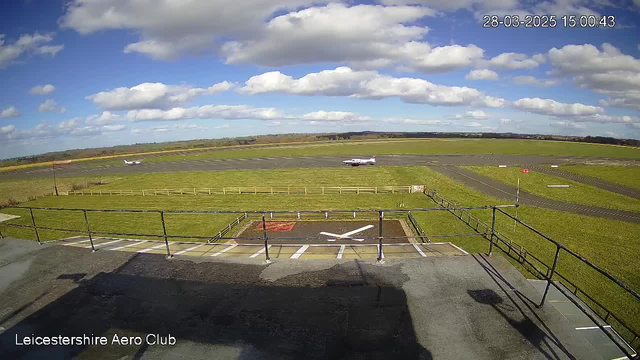 A view from a webcam positioned at the Leicestershire Aero Club, showing an airfield. The sky is mostly clear with scattered white clouds. In the foreground, there is a tarmac area with a helipad marked by a large white 'X' and a red square. Behind the helipad, the airstrip is visible, with a small plane taxiing on the runway. Green fields surround the airstrip, and wooden fencing borders the area. The time displayed on the image is 15:00:43 on March 28, 2025.