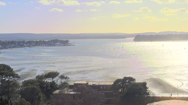 A scenic view of a shimmering body of water under a pastel sky, with soft clouds scattered above. In the foreground, trees frame the view, while a building is partially visible at the bottom. Further out on the water, colorful kites can be seen, suggesting sailing or water sports. In the distance, a shoreline with houses is visible, along with a gentle horizon line marked by distant hills. The overall atmosphere is tranquil, with light reflecting off the water's surface.