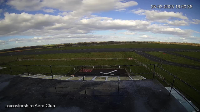 A view from a webcam at Leicestershire Aero Club. In the foreground, there is a tarmac area with a white cross marking the landing zone, surrounded by a low fence. Behind this are green fields that stretch towards the horizon. The sky is partly cloudy with patches of blue visible. The image features a mix of grass and asphalt surfaces, suggesting a rural aerodrome setting.