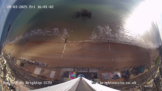 Aerial view of a coastline with a sandy beach and gentle waves in the foreground. A pier extends into the water, partially submerged, with remnants visible. The beach is dotted with people walking and lying on the sand, and colorful beach huts or tents are visible along the shoreline. In the lower part of the image, there are several structures and attractions, including a circular feature that may be a pool or ride. The scene is illuminated by bright sunlight reflecting off the water, creating a shimmering effect. The image also includes a timestamp and a watermark indicating it is powered by Brighton CCTV.