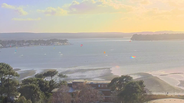 A scenic beach view featuring calm waters reflecting a sunset, with colorful kitesurfers and sailboats dotting the horizon. In the foreground, there are trees and buildings near the shore, with gentle waves lapping against the sandy beach. The sky has a mix of soft clouds and warm colors, creating a tranquil atmosphere.