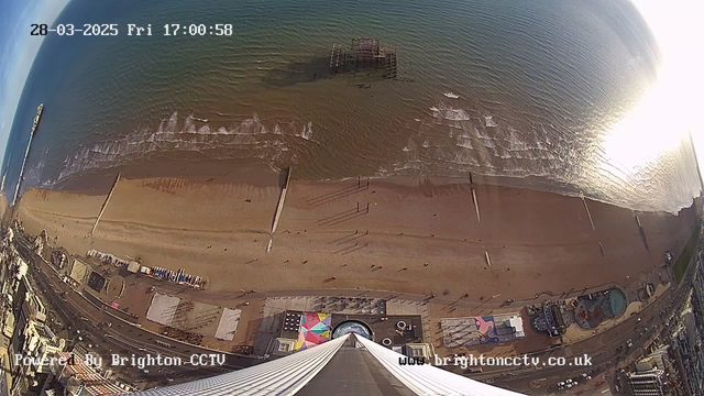 A high-angle view of a beach scene, showing golden sand meeting calm blue waters. Small waves ripple towards the shore. In the distance, a partially submerged pier is visible. The beach is dotted with a few people walking and lounging. Below, colorful structures and beach equipment are arranged, alongside a promenade. The image is timestamped for March 28, 2025, at 17:00.