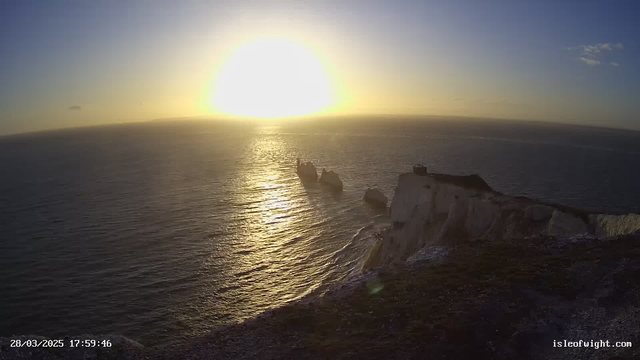 A scenic coastal view at sunset, with a glowing sun on the horizon shining over calm waters. The coastline features rocky formations, with some larger rocks jutting out of the sea. Gentle waves create a shimmering effect on the water's surface, and the sky transitions from blue to golden hues as the day ends. The rugged cliffs are visible in the foreground, with some grassy patches.
