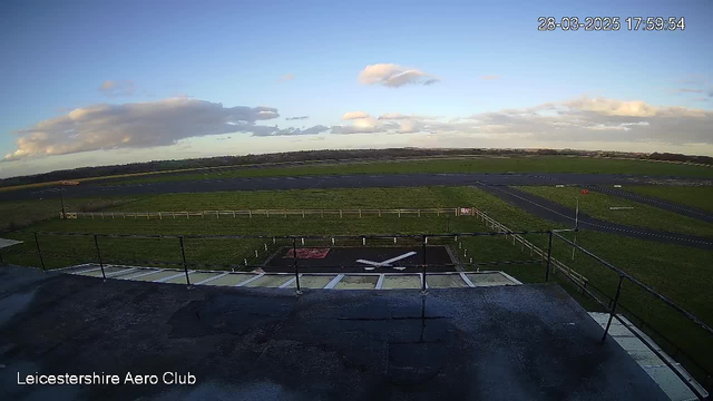 A clear sky with scattered clouds is visible in the background. In the foreground, there is a grassy field surrounded by a white fence. A section of an airstrip, marked with a large white cross, is in the center. To the right, there is a red sign. The image is taken from a higher vantage point, showing the landscape below, including the field and runway.