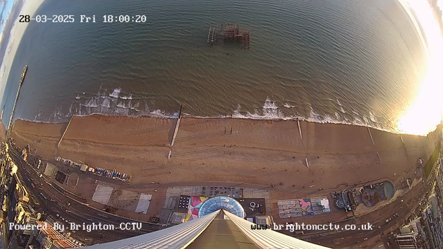 A high-angle view of a beach and ocean. The beach has golden sand with some people walking near the water’s edge. Gentle waves approach the shore, creating white frothy edges. An old pier structure is partially submerged in the water, visible in the mid-distance. To the bottom right, there are colorful beach attractions including a circular structure and several other features, along with a parking area. The image has a timestamp indicating the date and time at the top, and a logo and website URL at the bottom.