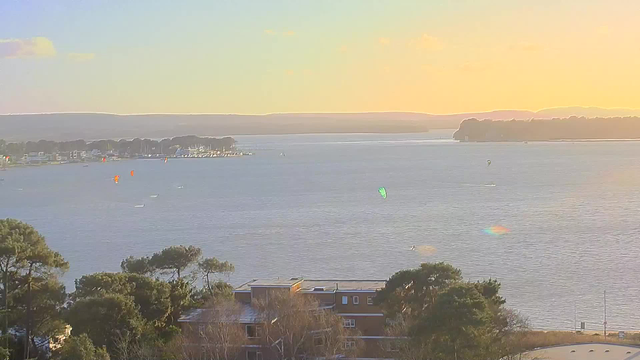 A scenic view of a calm body of water at sunset, with a soft orange and yellow sky. Silhouettes of trees and buildings are in the foreground, while several boats and kites can be seen on the water, including a green kite. In the distance, a shoreline with hills can be observed, adding depth to the landscape.