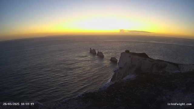 A scenic view from a coastline during sunset. The sky transitions from deep blue to warm orange and yellow hues near the horizon. Below, the calm sea reflects these colors. In the foreground, white cliffs rise sharply, with a rocky formation partially submerged in water. A tall structure is visible on the cliffs, adding a point of interest to the landscape. The scene conveys a tranquil and picturesque atmosphere.